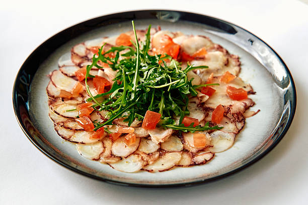 istock A beautifully arranged dish featuring thinly sliced octopus served with fresh greens and diced tomatoes, creating a delightful presentation of seafood cuisine. 2226925635