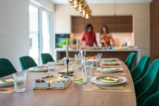An elegant dinner table with green chairs, set with plates, glasses, and decor in a bright room. Two women prepare food in a modern kitchen with warm lighting in the background.