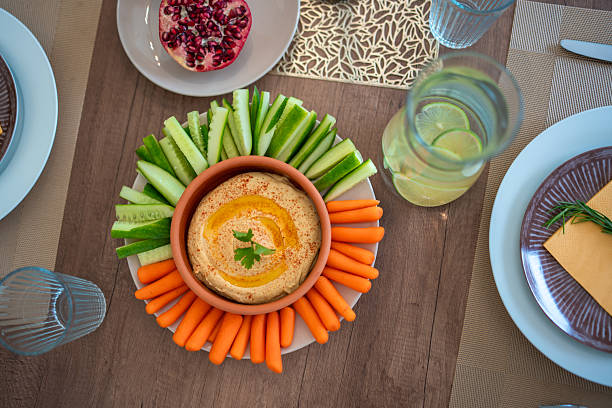 Top view of an elegant dinner table featuring a hummus platter garnished with paprika and parsley, fresh carrot and cucumber sticks, pomegranate seeds, and refreshing lemon water in a glass pitcher.