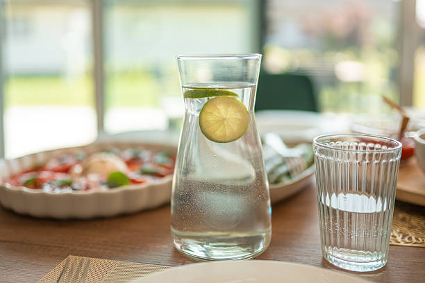 A well-lit indoor dining setup featuring a carafe of lime-infused water, a glass, and a vibrant fresh salad. Natural light streams in through large windows, creating a serene and inviting atmosphere for a meal.