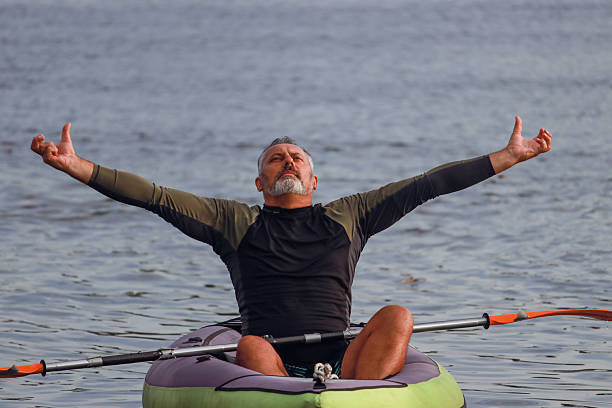 A joyful middle-aged man kayaking on calm water. He is seen paddling, stretching, and expressing happiness and freedom. The serene outdoor setting reflects a peaceful moment of recreation and adventure. Dressed in casual sportswear, he looks relaxed and content, celebrating nature and active lifestyle in middle age.