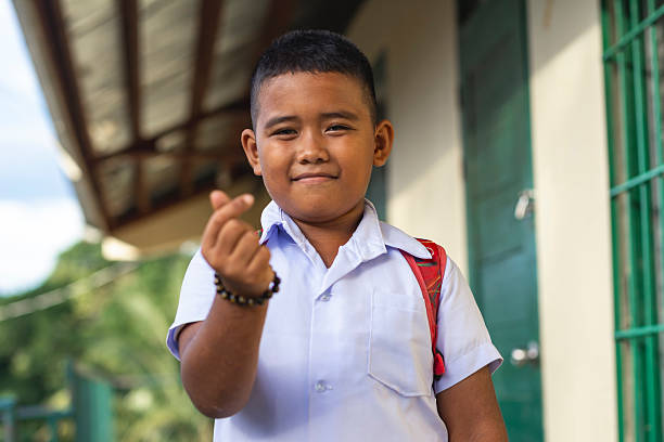 A poor but friendly rural school kid makes a finger heart gesture. Outside a simple small classroom building.
