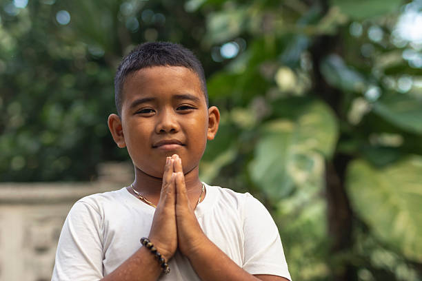A respectful young asian boy says please, with hands clasped. Asking for something in a polite manner. Outdoor garden scene.