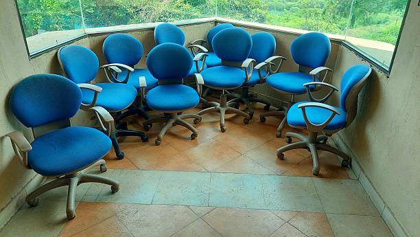 A cluster of blue upholstered office chairs with wheels is arranged in a tiled corner. The scene is set against a curved wall with large windows overlooking a lush green landscape. The chairs have rounded backs and seat cushions, with grey armrests and base. The flooring consists of square tiles in earthy tones, creating a warm atmosphere. The configuration suggests a casual or collaborative workspace.