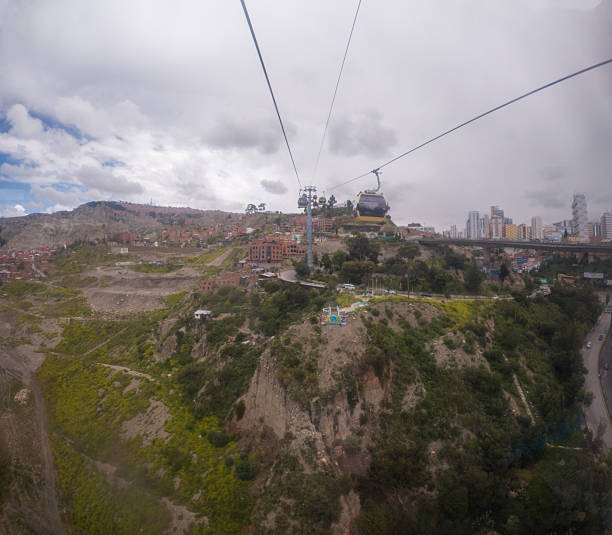 Cable cars traveling above green hills and rocky cliffs near a city with buildings and roads. Aerial view of urban hillside transportation and natural terrain. Cityscape with public transit system.