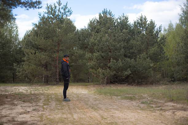 Man standing alone on a forest path, dressed in a black jacket and orange hoodie with hands in pockets, surrounded by tall green pine trees under a partly cloudy sky. Outdoor solitude concept.