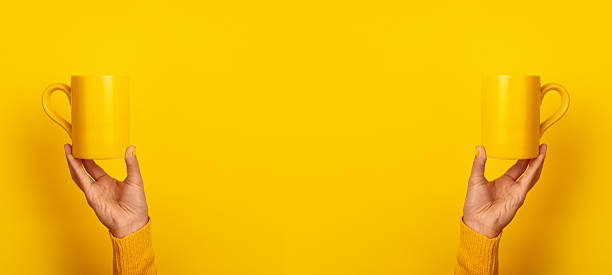istock Symmetrical image of two hands in yellow sweaters holding yellow ceramic mugs against a bright yellow background. 2226951738