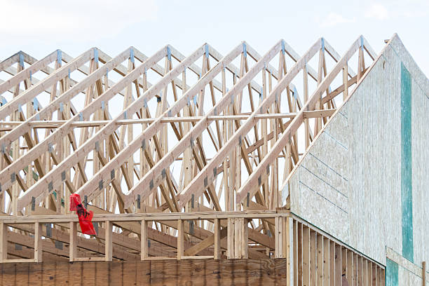 Close-up of wooden roof trusses with metal brackets in a house under construction, showcasing structural framework and carpentry against a clear blue sky.