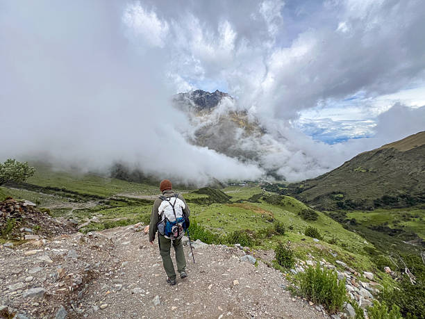 Man with backpack and trekking pole walking on rocky mountain path surrounded by green hills and low clouds. Outdoor adventure in misty highland landscape. Hiking and nature exploration.