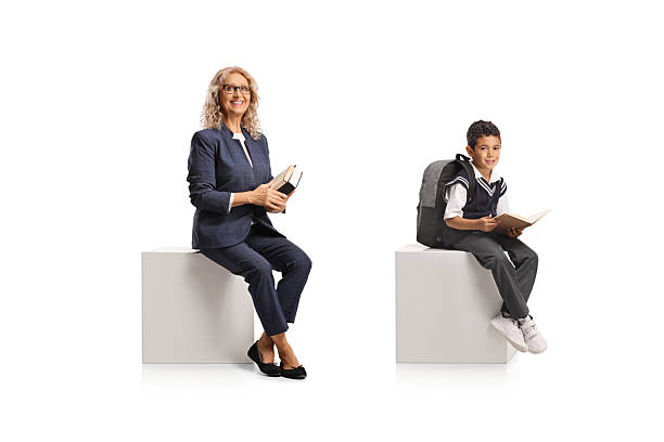 istock Boy in a school uniform holding a book and sitting on a white cube next to a teacher 2226958852
