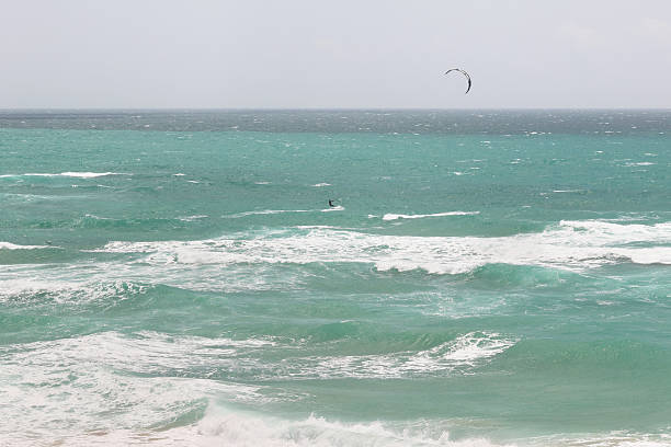 A man engages in kite surfing along the Mediterranean coast of Batroun, Lebanon. The sea is rough with strong waves and high wind, creating dynamic movement.