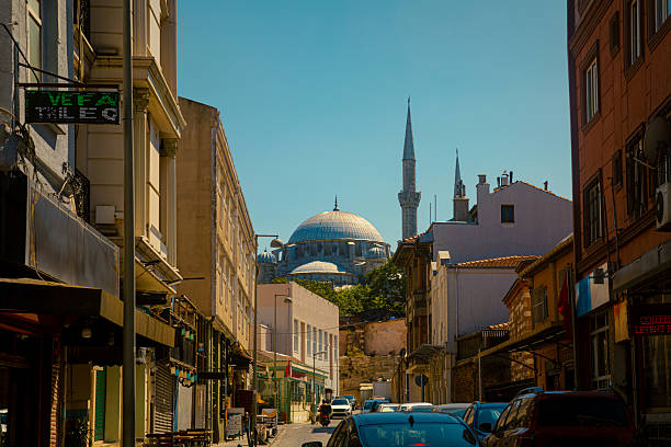Typical Istanbul street with colorful residential and commercial buildings, leading the eye toward the majestic silhouette of a grand Ottoman mosque. The minarets and domes rise elegantly in the background, capturing the harmony between daily urban life and centuries-old Islamic architecture. This image evokes a vibrant yet peaceful mood, ideal for themes of travel, heritage, Islamic culture, street photography, and timeless contrasts between tradition and modernity.