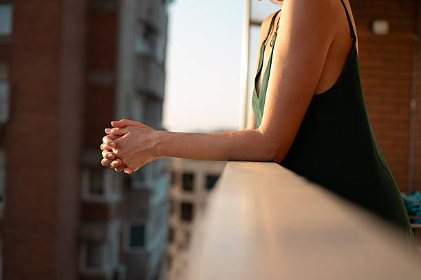 Close-up of woman leaning on balcony railing at sunset, enjoying a moment of relaxation. Soft light, urban background, peaceful mood.