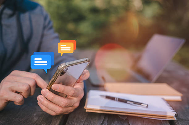 istock Social media marketing concept. Man Using Smartphone Outdoors typing, chatting conversation in chat box icons pop up with Notebook, Pen, and Laptop on Wooden Table with green park background 2226968695