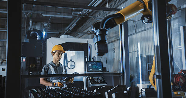 istock Young Asian Male Technician in a Yellow Hard Hat Operates a Robotic Arm with a Tablet Computer Software in an Advanced Manufacturing Plant. Metal Industry Specialist Performing Tasks at Work 2226975351
