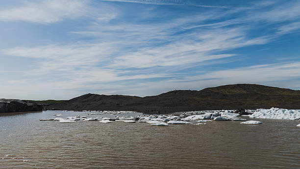 A serene glacial lagoon in Iceland with floating ice chunks on brownish water, rugged hills with green patches, and a partly cloudy blue sky.