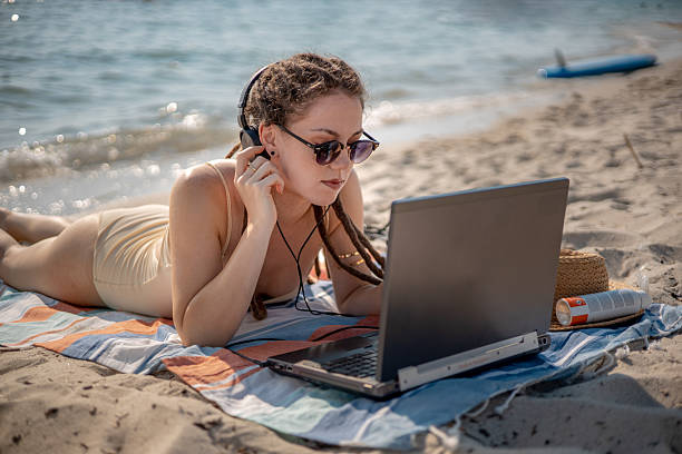 istock Remote female content creator with dreadlocks typing on her laptop while lounging on a beach mat, headphones on, enjoying freelance writing work by the ocean 2226999100
