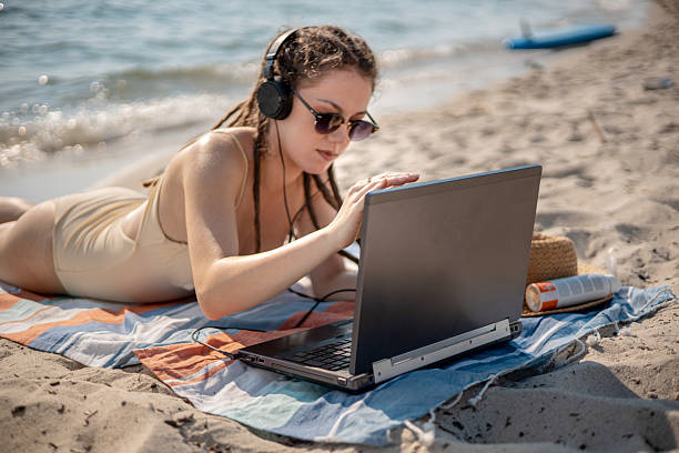istock Female web developer with dreadlocks working remotely on her laptop, lying on a beach mat with headphones, coding by the sea during summer 2226999101