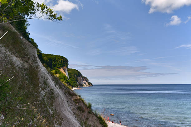 White chalk cliffs near KÃ¶nigsstuhl on Rugen island, Germany. Forested coast meets calm Baltic Sea under a warm summer sky with toursist bathing at beach.