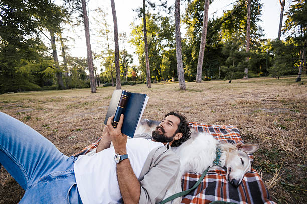 istock A man lying on a blanket outdoors, relaxing with his Russian Borzoi dog while reading a book. The peaceful scene captures a quiet moment of leisure and companionship in a natural setting. 2227006593