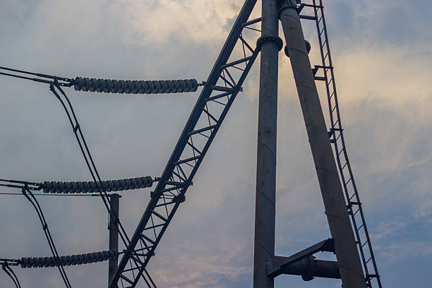 A low-angle shot of a high-voltage electricity pylon against a cloudy sky. The image shows the intricate network of cables, insulators, and metal framework of the pylon.