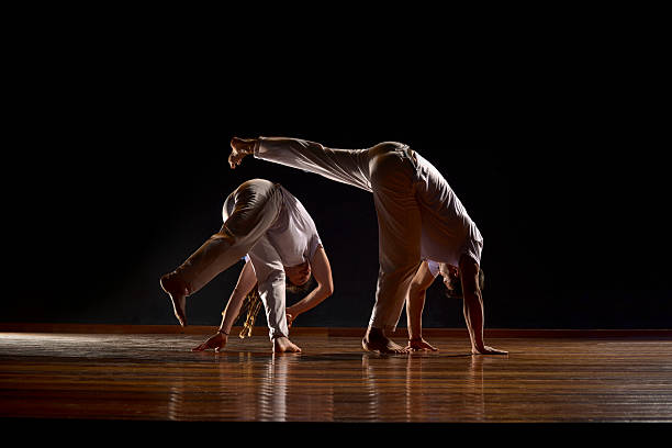 Two silhouetted athletes practice capoeira against a dramatic backlight. The image captures the energy and fluidity of the game (\