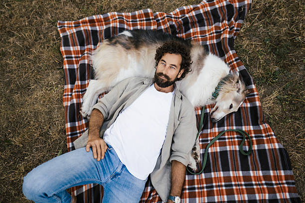 A man is lying on a checkered picnic blanket in a peaceful outdoor setting, while relaxing with his Borzoi dog.