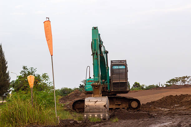 A large green construction excavator sits idle on a dirt construction site under a cloudy sky. The heavy machinery stands near mounds of dirt, with a windsock and trees in the background