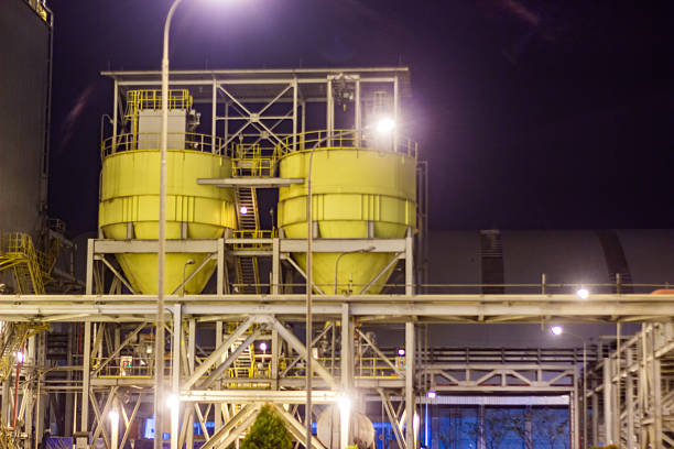 This image showcases an industrial plant at night, illuminated with bright lights. Yellow silos and metal structures are prominent, highlighting the complexity of modern manufacturing and industrial