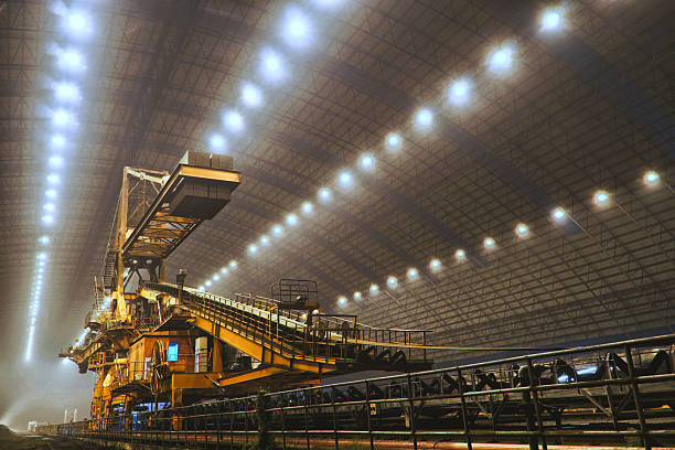 Captured in a spacious industrial setting, a colossal excavator stands prominently beneath a gracefully arched ceiling illuminated by rows of lights.