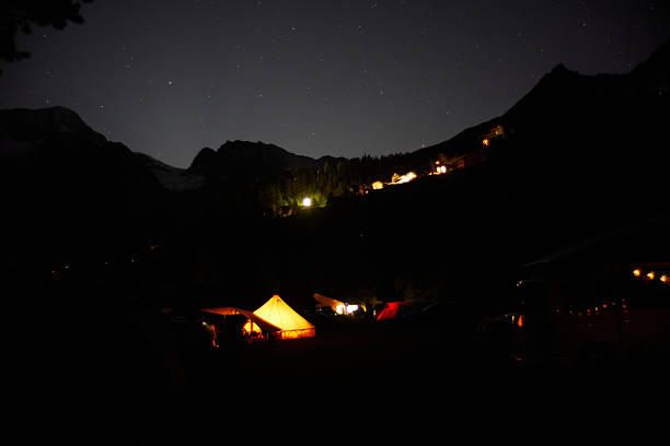 A glowing campsite with tents lit by lights under a dark, starry sky surrounded by mountains. Captures serenity and the beauty of night outdoors.
