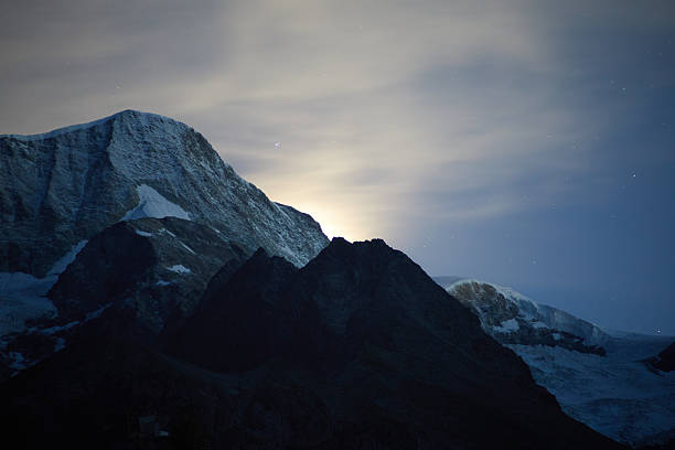 Mountains with snow-covered peaks glow subtly due to moonlight amid a tranquil and star-filled night, creating a serene and mystical atmosphere.