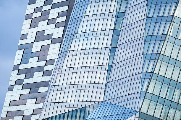 A contemporary building that showcases two contrasting facade styles. One section of the structure is curved and covered in reflective glass panels, creating a sleek and airy appearance with dynamic reflections of the sky. The other section is composed of bold black and white rectangular cladding arranged in an irregular geometric pattern, adding texture and visual depth. The combination of smooth glass and structured paneling highlights innovation in architectural design and material usage. Perfect for commercial use in construction presentations, real estate marketing, architectural studies, and editorial content focused on modern urban development.