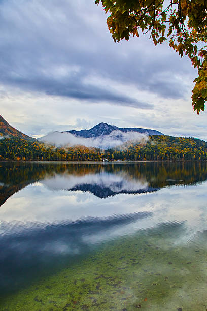 A serene lake reflecting mountains and surrounded by autumn foliage, under moody clouds. Perfect depiction of natural tranquility and vibrant fall colors.