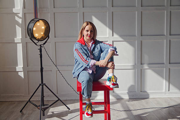 A Young Woman Sitting on a Red Stool in a Sunlit Studio Wearing a Colorful Denim Outfit, Surrounded by Industrial Lighting Equipment and Casting Soft Shadows on a White Paneled Wall Background