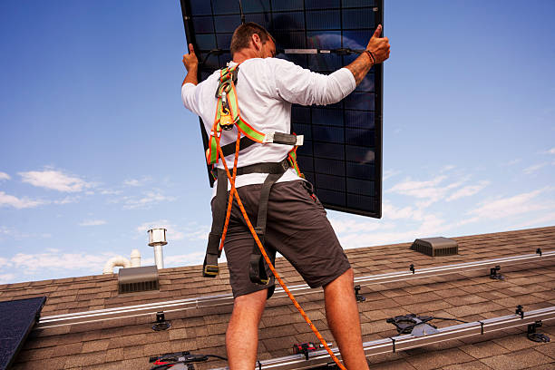 A male worker in a safety harness lifts a solar panel into position on a residential roof under a clear blue sky. His gear ensures fall protection while he participates in clean energy implementation, showcasing both occupational safety and progress toward sustainable energy solutions.