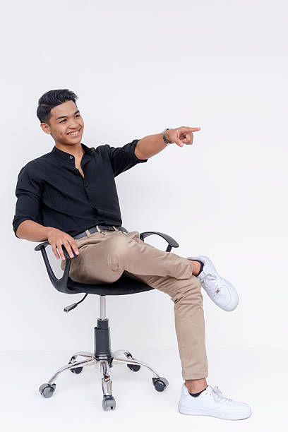 istock A optimistic and upbeat young man pointing to something at the right while sitting crossed legged on an office chair. Studio shot isolated on a white background. 2227032312