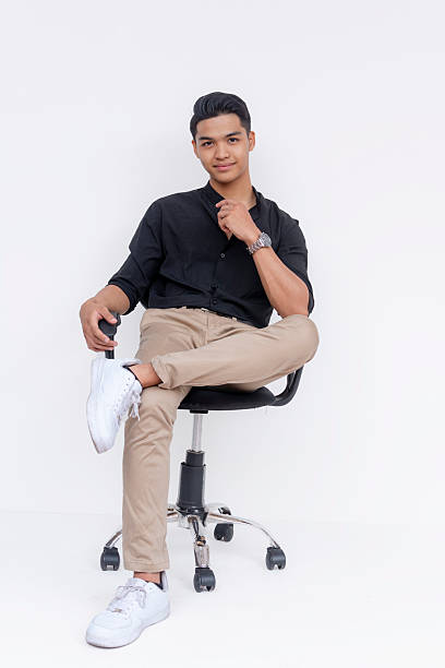 A confident and handsome Filipino man sitting relaxed, leg crossed, on an office chair. Studio shot isolated on a white background.