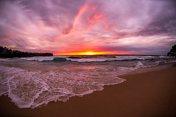 A dramatic sunrise lights up the sky over the shoreline in Sydney, Australia, with glowing clouds, crashing waves, and golden reflections on the sand.