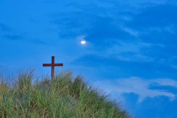 Wood cross on a New Zealand sand dune hill, with storm clouds on the horizon. Also the the moon shining bright.