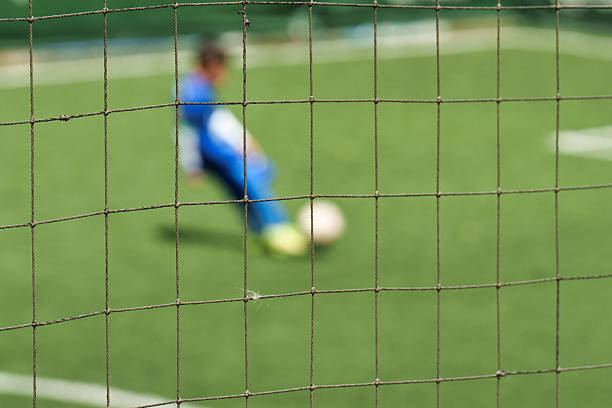 Close-up football goal net, seeing a young boy soccer goalkeeper watching his teammate playing. Youth Soccer game on a sunny summer school tournament Day. A football match going on in a background in Bangkok, Thailand.