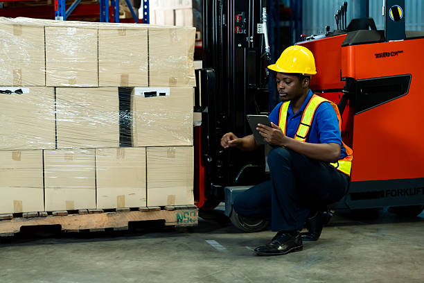 istock African male warehouse staff sitting on floor near boxes using tablet for checking stock data beside forklift inside storage facility, inside logistics warehouse. 2227045983