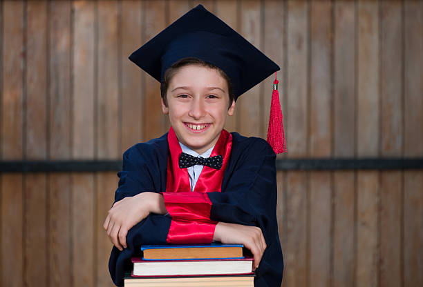A cheerful male elementary school student in a graduation outfit smiles as he leans against a stack of colorful books in front of a wooden wall