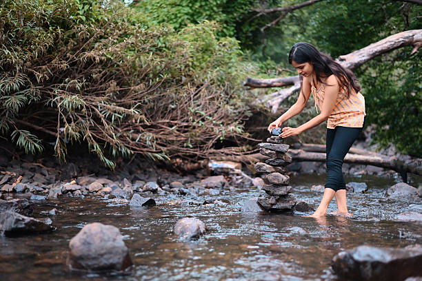 A joyful young woman carefully places a stone on top of a balanced rock stack in a shallow forest stream. Surrounded by lush greenery and fallen trees, she enjoys a peaceful moment of mindfulness and creativity in nature. Balance, meditation, nature therapy, outdoor leisure, and eco-friendly travel.