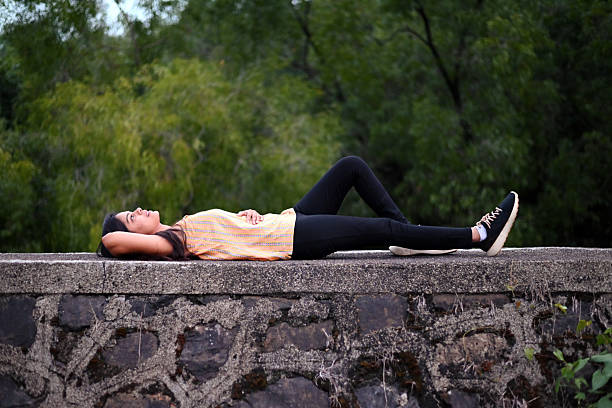 A young woman lies comfortably on her back atop a stone wall in a natural outdoor setting. She appears peaceful and relaxed. Surrounded by lush greenery, the scene conveys a mood of calmness, solitude, and connection with nature. Mental wellness, relaxation, freedom, and mindful living.