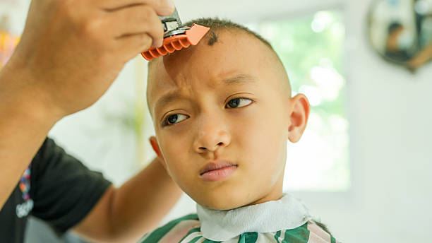 Young boy with shaved head is getting haircut bright, modern barbershop. barber is using clippers to shape boy hair, while boy looks thoughtfully camera, showcasing mix of curiosity