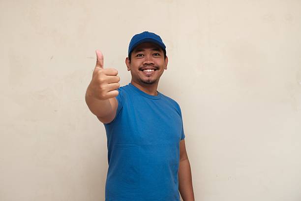 istock Cheerful man in blue shirt and cap smiling while giving a thumbs up, expressing approval, success, or positive feedback. 2227075865