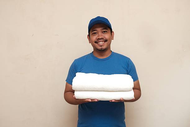 Cheerful man in blue shirt and cap holding folded white towels, representing cleanliness, hospitality, or laundry service.