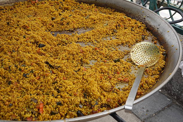 Large paella pan cooking outdoors, filled with vibrant saffron rice, fresh vegetables, and a gleaming serving spoon ready for sharing