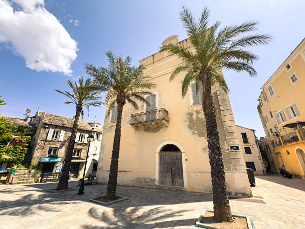 Old yellow building with wooden door and balcony framed by tall palm trees. Sunny Mediterranean village square with stone pavement and traditional Corsican architecture. Coastal town ambiance.